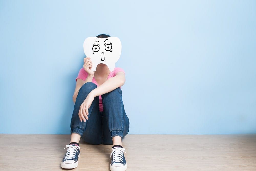 Woman holding a paper cut out of a giant scared tooth in front of her face while sitting against a light blue background